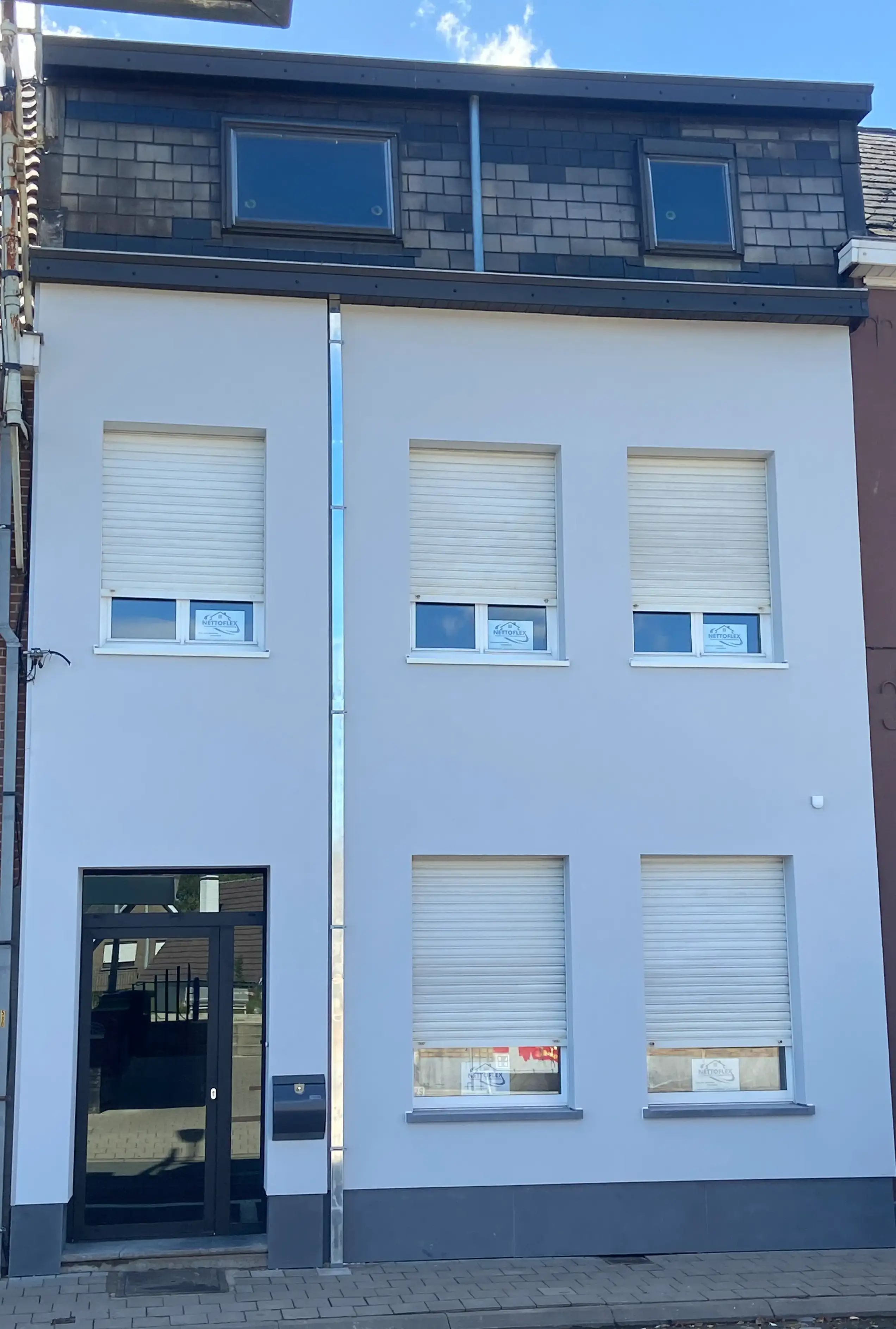 Woninggevel met lichtgrijze crepi, witte rolluiken en donkergrijze dakopbouw met twee dakramen.
