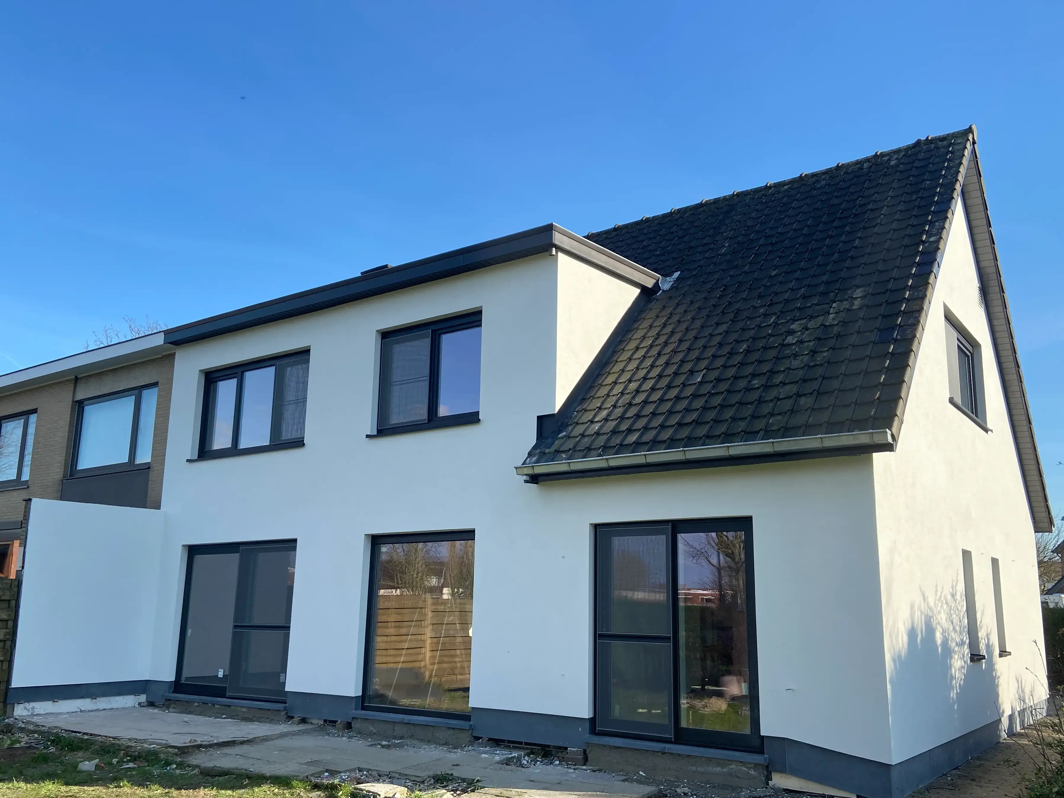 Achterzijde van een woning met witte crepi-gevel, donkere ramen en schuifdeuren naar de tuin, onder een donker getiled zadeldak.