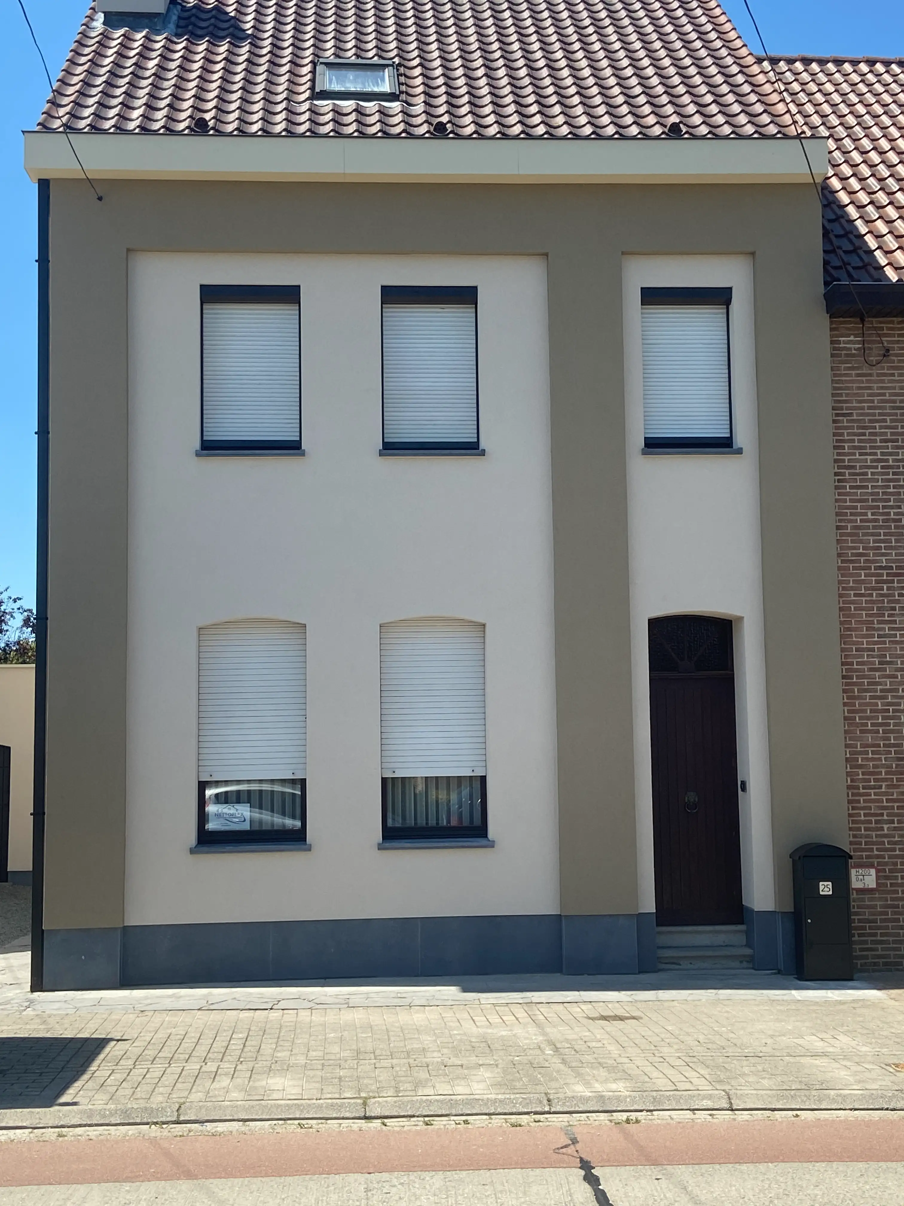 Twee verdiepingen hoge woning met beige crepi-gevel, ramen met witte rolluiken, donkere houten voordeur en rood pannendak met dakraam.