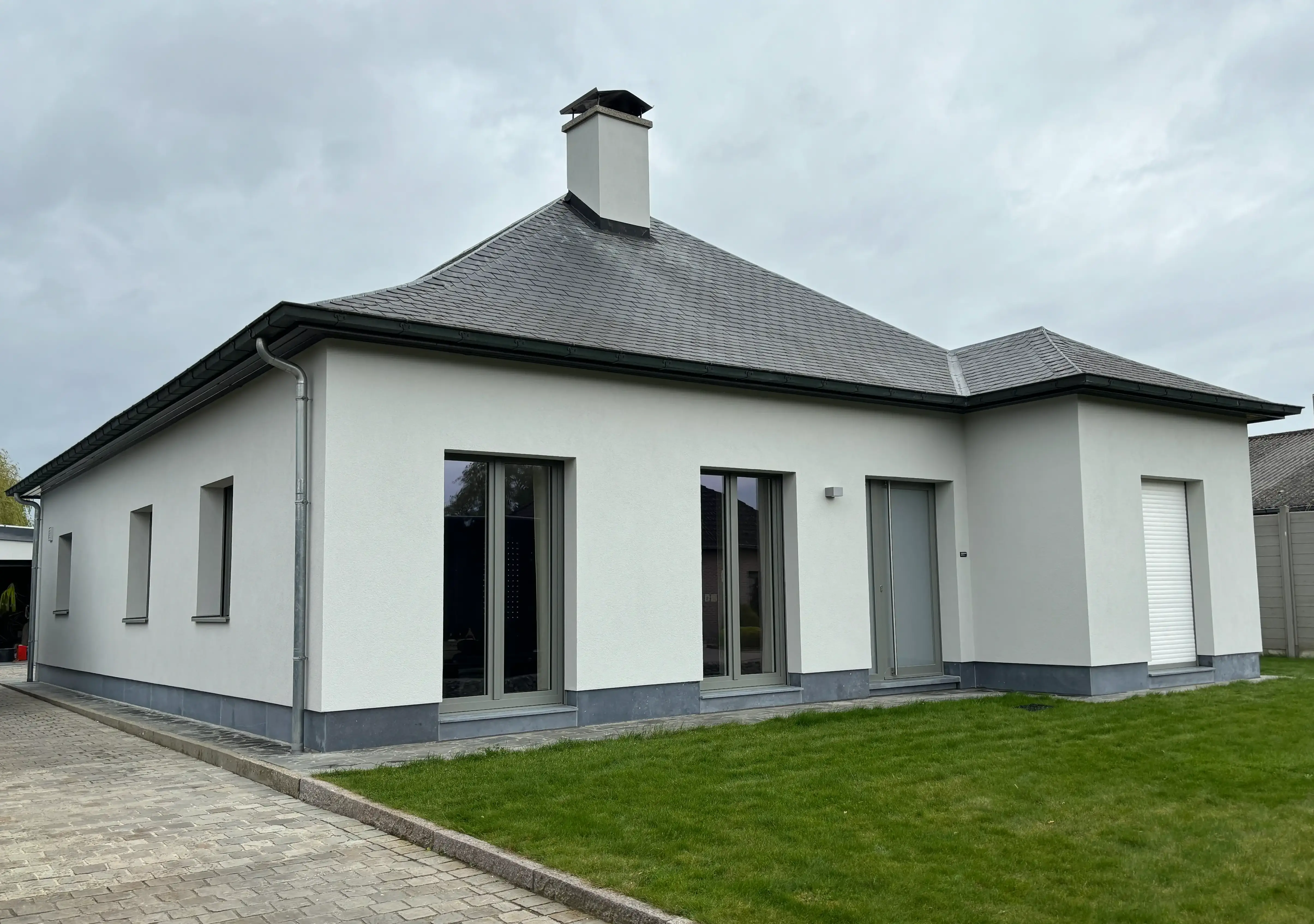 Woning met lichtgrijze crepi-gevel, grote glasdeuren, antracietkleurig zadeldak en groen gazon.