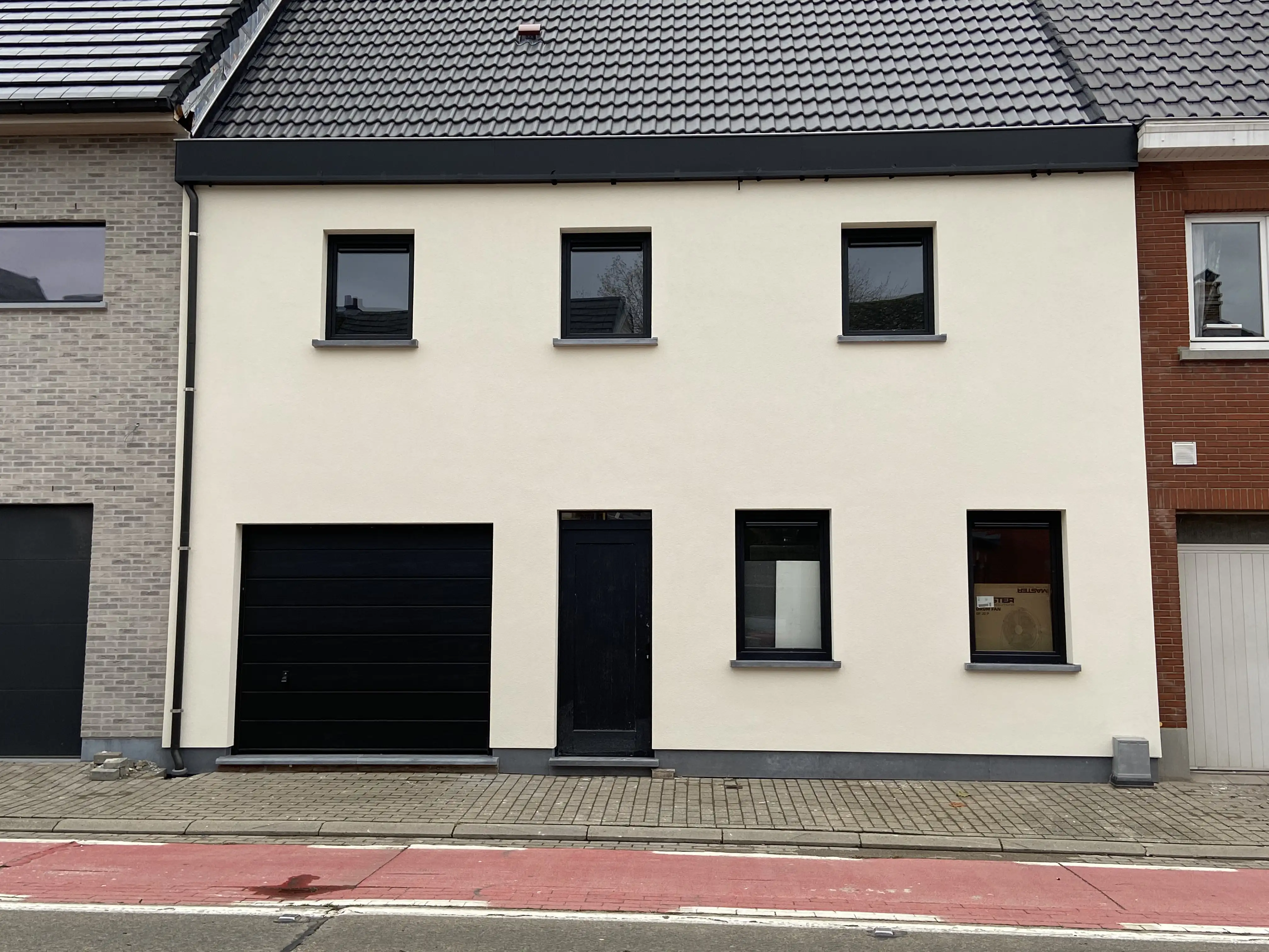 Twee verdiepingen hoge woning met crèmekleurige crepi, zwarte ramen, zwarte garagedeur en donker getiled zadeldak.
