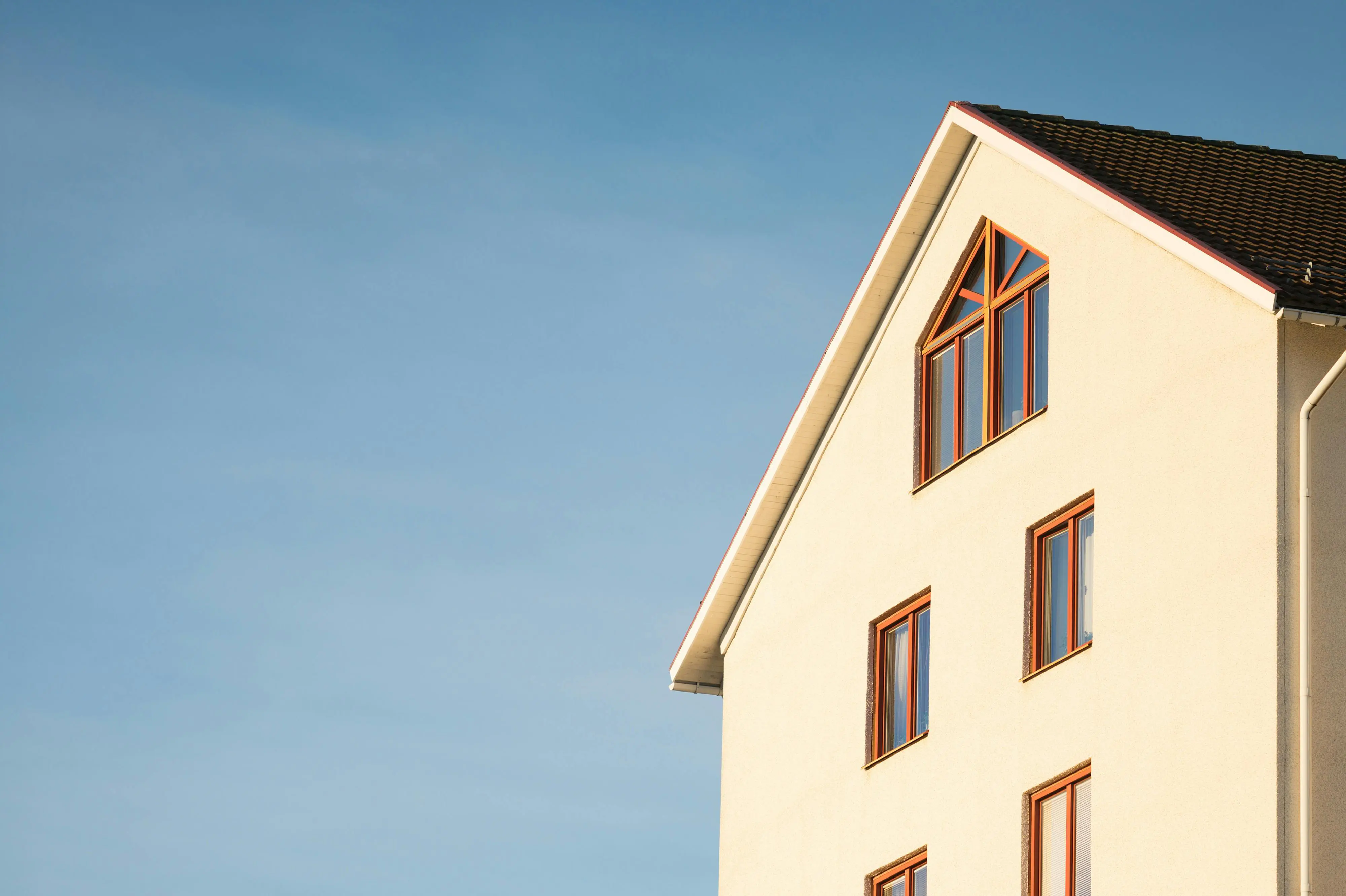 Gedeelte van een meerlaagse woning met beige crepi-gevel, houten kozijnen en donker getiled zadeldak tegen blauwe lucht.